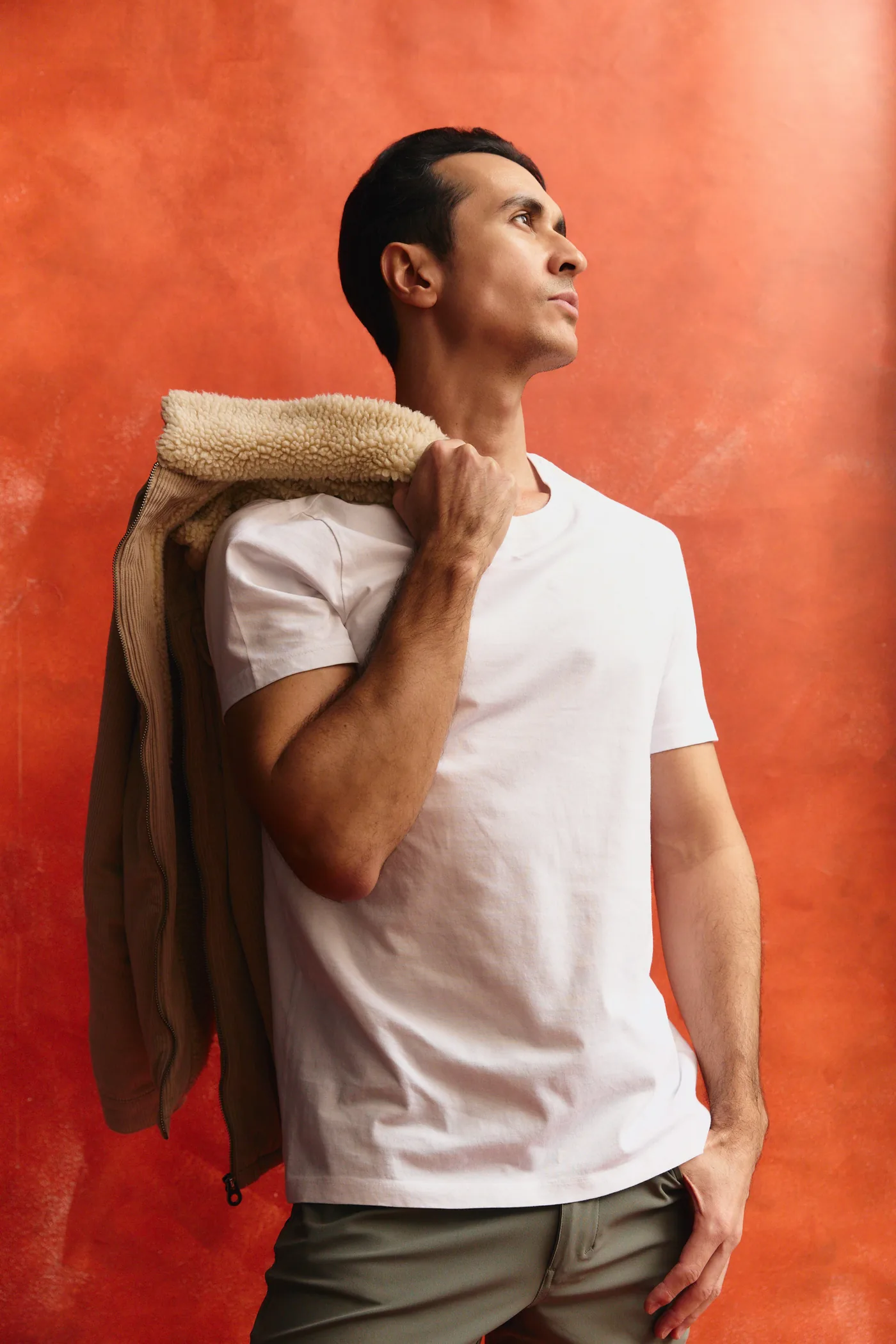A man in a white T-shirt holding a tan jacket over his shoulder against an orange wall