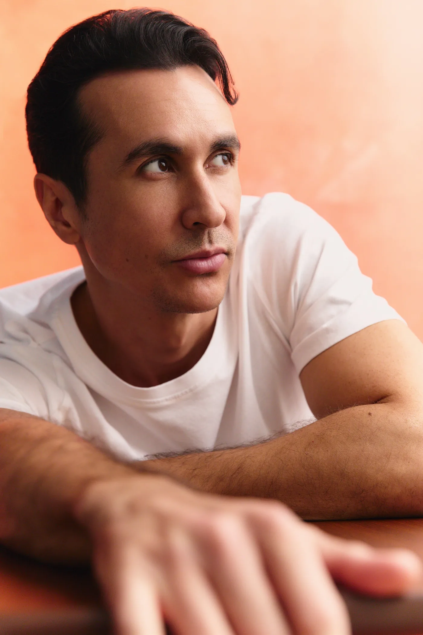 A man in a white heavyweight T-shirt leaning against a peach wall