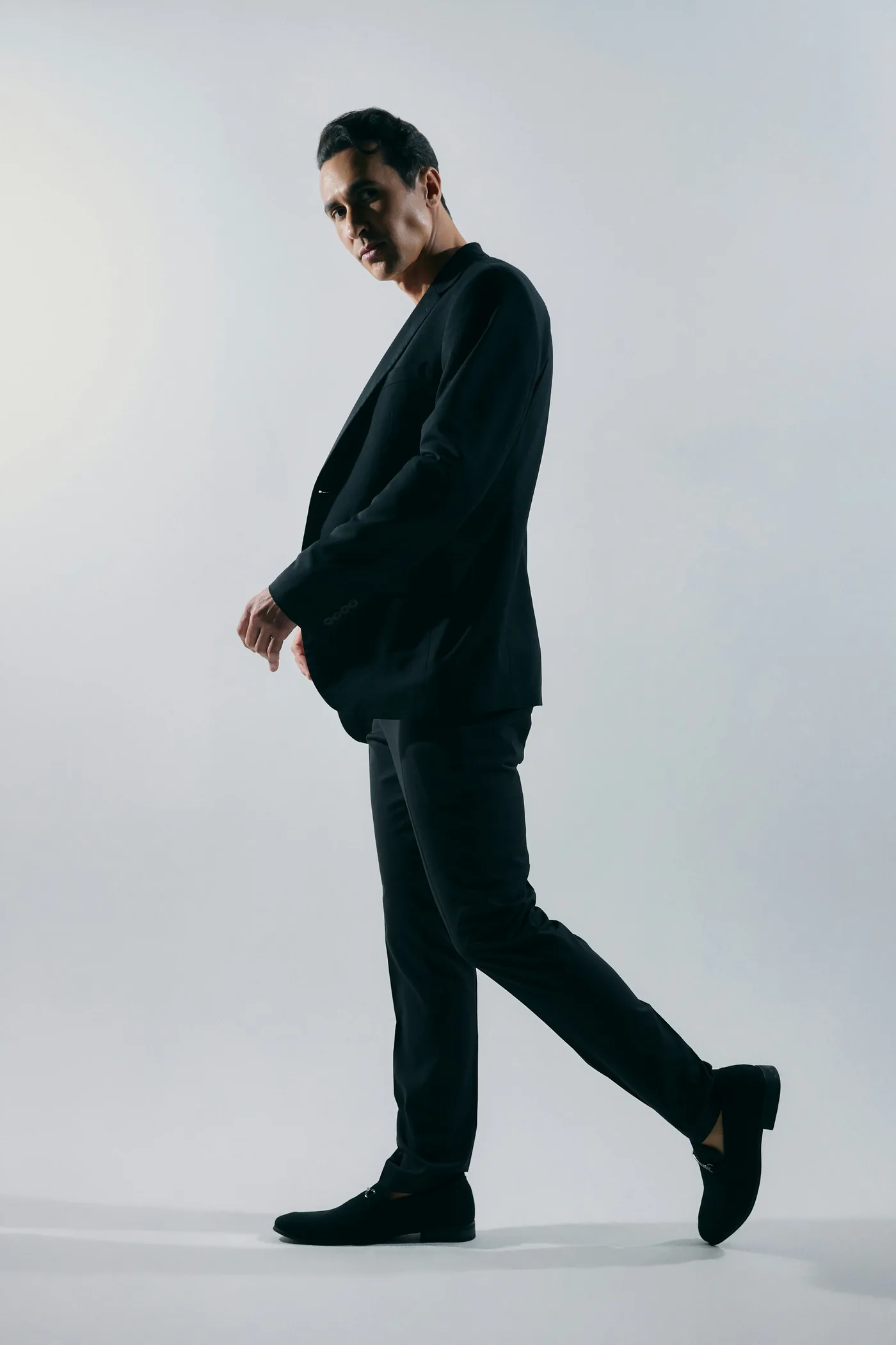A man in a black suit walking against a pale grey backdrop